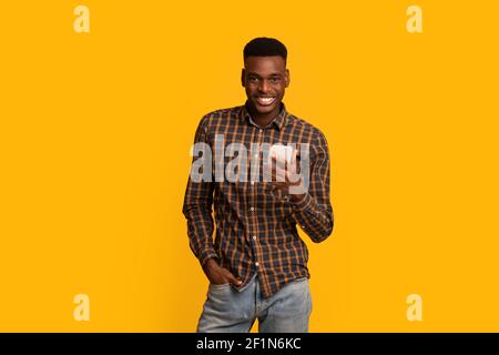 Portraif di bel giovane afroamericano Guy con smartphone in mani che posano su sfondo giallo. Allegro Black Millennial Man con cellulare e Foto Stock