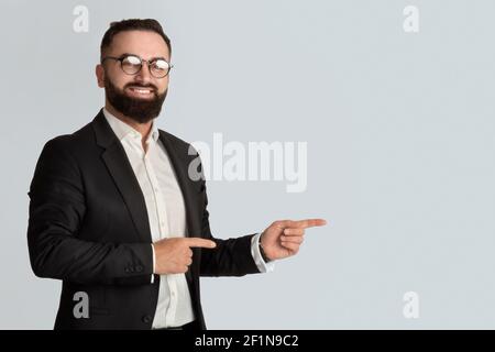 Uomo d'affari serio in ufficio indossare tand occhiali sorridenti e puntando da parte su sfondo grigio chiaro, spazio per voi annuncio. Prom. Manager con portatore millenario Foto Stock