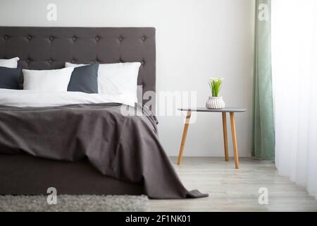 Stile moderno e design alla moda negli interni degli appartamenti. Letto per due con coperta e cuscini, pianta in vaso su tavolo su sfondo grigio parete, grande Foto Stock