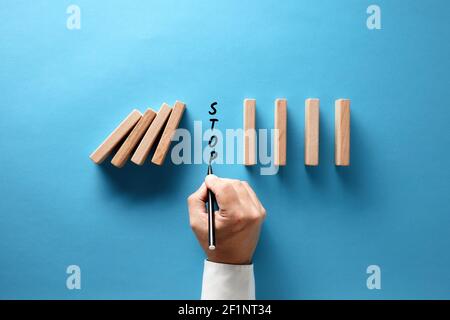 Mano di uomo d'affari che scrive la parola stop per impedire che i dominos di legno collassino su sfondo blu. Foto Stock