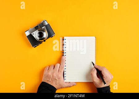 La mano maschile tiene una penna su un notebook vuoto o vuoto accanto a una vecchia fotocamera vintage su sfondo giallo. Foto Stock