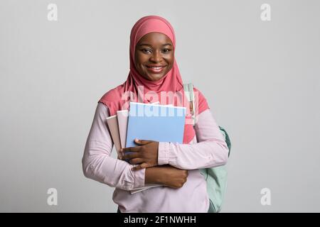 Bella africana americana studentessa giovane donna in hijab in posa con notebook e libri su sfondo grigio studio, trasportando zaino, allegro Foto Stock