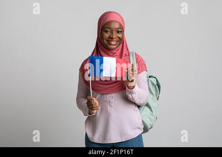 Donna nera positiva in hijab mostrando bandiera della Francia, con zaino sulla spalla, sfondo grigio studio, spazio copia. Sorridente musl afroamericano Foto Stock