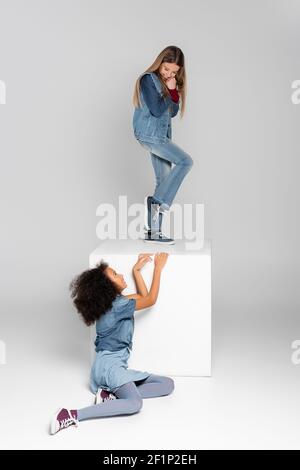 ragazza allegra in piedi su cubo bianco mentre afroamericana ragazza seduto su grigio Foto Stock