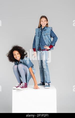 bambini interrazziali in abiti eleganti in denim che si posano su scatola bianca isolato su grigio Foto Stock