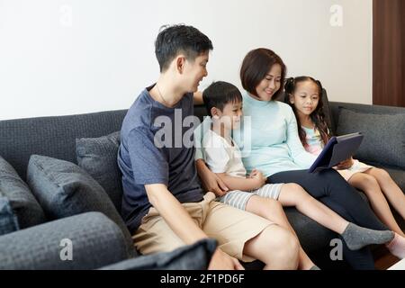 Felice famiglia vietnamita con due bambini che si divertono a stare insieme e guardare video animati o blog su tablet digitale Foto Stock