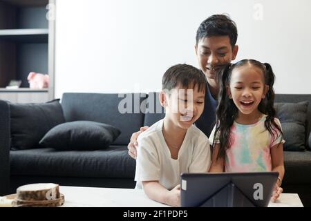 Felice padre ridente, suo figlio e sua figlia che guardano un simpatico cartone animato o uno spettacolo su un tablet digitale Foto Stock
