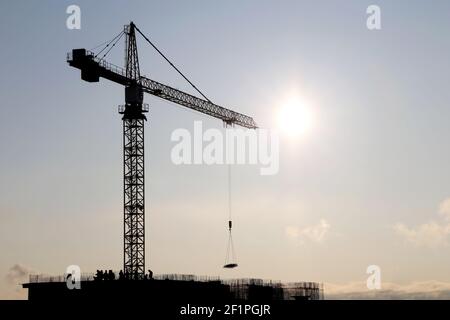 Silhouette di gru da costruzione e lavoratori in un edificio residenziale incompiuto contro il sole. Costruzione di alloggi, condominio in città Foto Stock