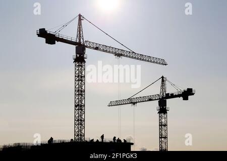 Sagome di gru e operai edili su edifici residenziali incompleti contro il cielo e il sole splendente. Costruzione dell'alloggiamento Foto Stock