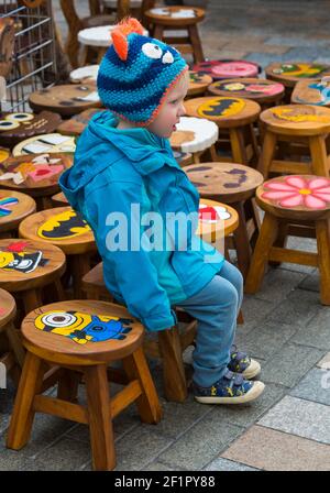 Giovane ragazzo seduto su sgabelli in legno decorati dai colori vivaci al Gloucester Quays Victorian Christmas Market, Gloucester, Gloucestershire nel mese di novembre Foto Stock