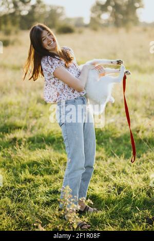 Elegante donna felice che gioca con simpatico cucciolo bianco in calda luce del tramonto nel prato estivo. Casual giovane femmina ridere e tenere pazzi attivi svizzeri Foto Stock