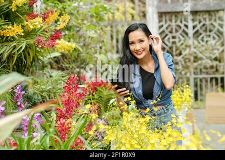 Felice giovane donna asiatica fotografare piante in fiore cameretta e sorridente alla macchina fotografica Foto Stock