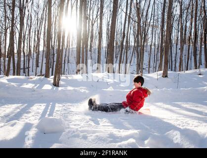 Un bambino che dorme giù su una collina innevata in una zona boscosa durante il giorno di sole. Foto Stock