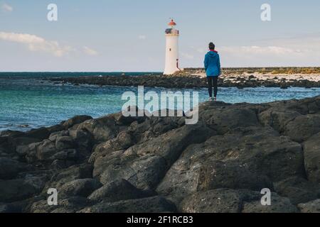 Giovane donna che guarda un faro in lontananza in cima alla costa rocciosa, Port Fairy, Australia. Foto Stock