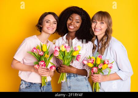 Foto di bella ottimista tre Signore tenere fiori indossare bianco camicia isolata su sfondo giallo brillante Foto Stock