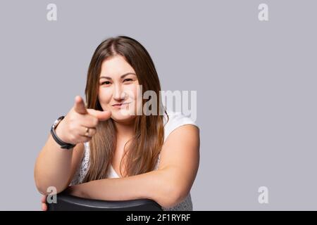 Ritratto di una donna chubby che pone e punta il suo dito indice verso la fotocamera. Sfondo grigio con spazio laterale vuoto. Foto Stock