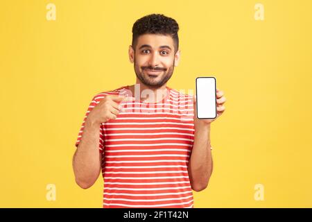 Uomo sicuro e positivo con barba in t-shirt a righe che punta il dito sullo smartphone con display bianco vuoto, indossarlo con l'applicazione. Monolocale interno Foto Stock