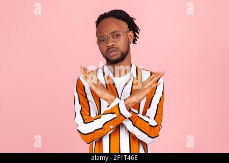 Questo è il fine. Interessato serio uomo africano con Dreadlock in occhiali ed elegante camicia luminosa che attraversa le mani mostrando x segno, rifiuto. Interno Foto Stock