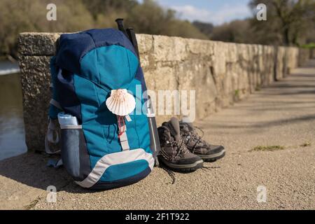 Zaino con simbolo di Camino de Santiago, scarponi da trekking e pali appoggiati su un muro di pietra. Pellegrinaggio a Santiago de Compostela. Spazio di copia Foto Stock
