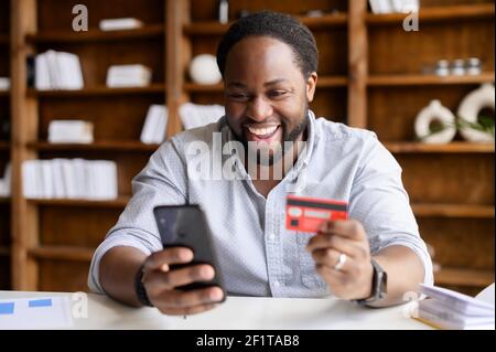 Eccitato afroamericano ragazzo sta usando uno smartphone e una carta di credito per lo shopping online, felice uomo nero sta ordinando cibo online, maschio pagare per l'acquisto a lungo atteso sul telefono cellulare. E-banking Foto Stock