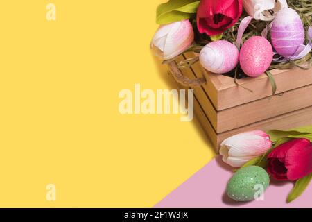 Uova colorate di Pasqua in un cestino e tulipani su uno sfondo giallo illuminante alla moda. Concetto di pasqua minimo. Carta di Pasqua con spazio per la copia del testo. Foto Stock