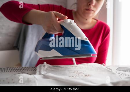 Donna caucasica che stira i vestiti mentre si siede a casa in luminoso camera - persone reali attività domestiche concetto primo piano stira in mano Foto Stock