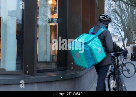 Il pilota Deliveroo con zaino attende nella coda di ritiro all'esterno del ristorante per i piatti da asporto dell'ordine del cliente Foto Stock