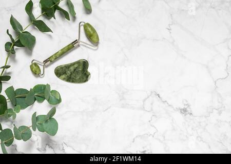 Rullo di giada verde e pietra di gua sha per il massaggio del viso e ramo di eucalipto su sfondo marmoreo. Accessori per la bellezza e l'autosocura, rullo per il viso Foto Stock