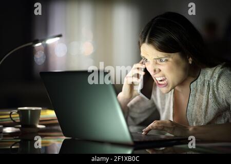 Freelancer arrabbiato che parla sul telefono cellulare che controlla il computer portatile nel notte in ufficio Foto Stock