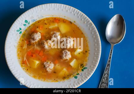 Zuppa con polpette in un piatto bianco di ceramica e un cucchiaio di metallo su blu. Porzione Pronta per mangiare la zuppa. Polpette, patate, carote, cipolle, pasta, pomodoro Foto Stock