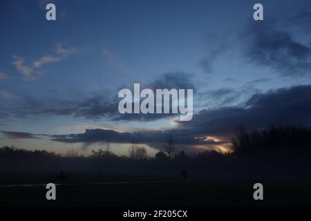 alba arancione con alberi silhouette in lontananza con un cielo blu e nuvole illuminate dal sole Foto Stock