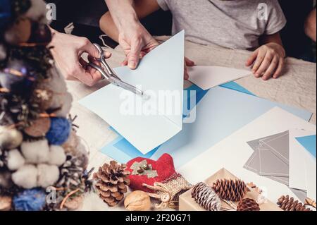 Le mani di una madre caucasica e di sua figlia stanno tagliando i fiocchi di neve di carta con le forbici. Fatti a mano, decorazioni natalizie, attività invernale familiare. Clos Foto Stock