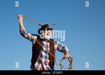 Cowboy occidentale senior che lancia la corda di lazo. Uomo selvatico bearded occidentale con giacca marrone e cappello che cattura cavallo o mucca. Rodeo o ranch. Foto Stock