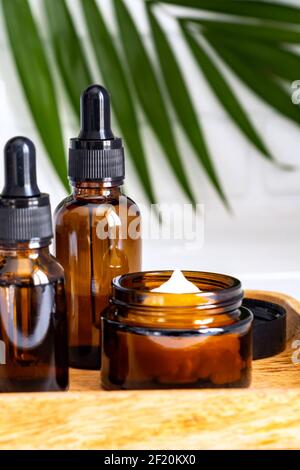 Bottiglie cosmetiche in vetro scuro con siero facciale o olio essenzioso e crema in un vassoio di legno con foglia di palma. Prodotti cosmetici di bellezza senza bradned Foto Stock