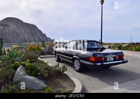 FRESNO, STATI UNITI - Mar 02, 2021: Una bella foto di una brillante auto Mercedes 560 SL nera parcheggiata di fronte Morro Bay Rock circondata da una pianta verde Foto Stock