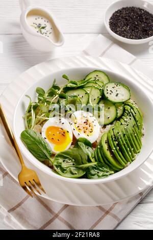 Ciotola Quinoa all'uovo con micrograni di girasole, cetriolo, avocado, spinaci in una ciotola bianca su un tavolo di legno bianco, vista verticale, cui turco Foto Stock