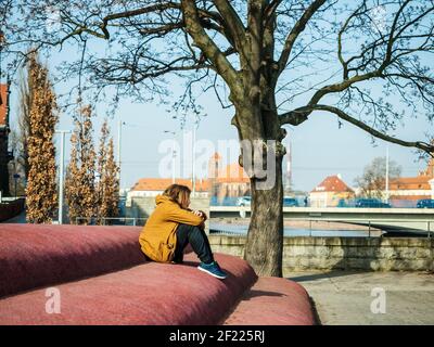 Triste giovane seduto da solo in una città, nello spazio urbano e guardando da un lato Foto Stock