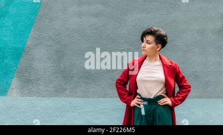 Ritratto candito giovane Brunette donna con capelli corti su fondo parete blu. Ritratto esterno donna moderna gen z in cappotto rosso Foto Stock