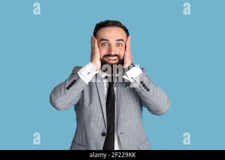 Ritratto di giovane uomo d'affari che tiene la testa in eccitazione, non può credere alla sua fortuna o successo su sfondo blu Foto Stock