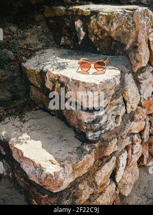 Paio di occhiali da sole arancioni seduti su gradini di pietra che conducono in basso ad una spiaggia con il sole che splende su di loro Foto Stock