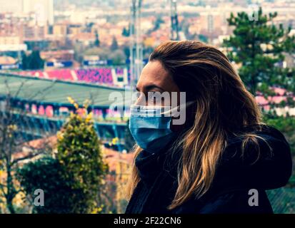 donna con maschera sulla vista del profilo del viso preoccupato per il coronavirus pandemia con vista di una città e stadio nel sfondo Foto Stock