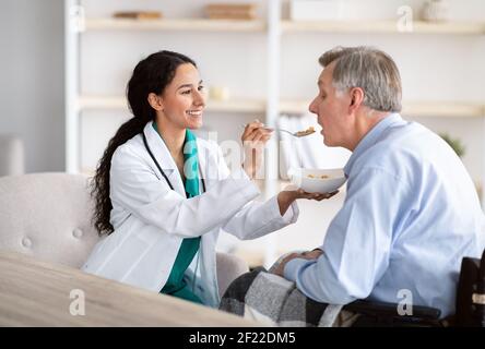 Assistenza medica per anziani con disabilità. Infermiera che alimenta l'uomo anziano handicappato in sedia a rotelle a casa Foto Stock