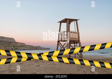 Nastro barriera - quarantena, concetto di isolamento, divieto di ingresso. Non incrociare. Una torre di bagnino sulla spiaggia mediterranea al tramonto durante l'estate. Foto Stock