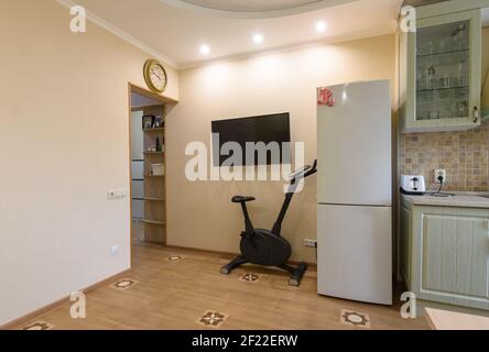 Un frammento dell'interno del soggiorno combinato con la cucina, una TV appeso alla parete, una cyclette e un frigorifero sono situati nelle vicinanze Foto Stock
