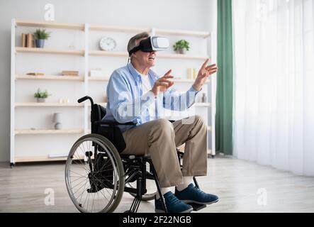 Felice uomo anziano in sedia a rotelle con disabilità che utilizza il visore VR per esplora la realtà aumentata a casa tua Foto Stock