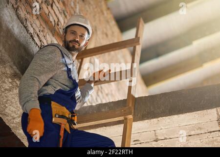 Bell'uomo da lavoro in tute blu e elmetto che guarda la macchina fotografica, scendendo lungo la scala mentre si lavora in cantiere. Costruzione, persone, concetto di professione Foto Stock