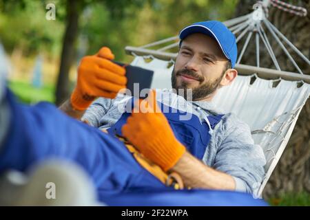 Felice giovane costruttore maschile indossare uniforme prendere una pausa, sdraiato in un amaca all'aperto e guardare qualcosa utilizzando smartphone in una giornata di sole. Costruzione, professione, concetto di riposo Foto Stock