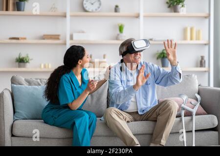 Uomo anziano disabile che utilizza il visore VR con un'infermiera giovane, esplorando la realtà aumentata a casa Foto Stock