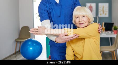 Artrite di un giunto a gomito in una paziente anziana in un centro di riabilitazione. Chiropractor aiuta a eseguire esercizi di recupero della salute della mano Foto Stock