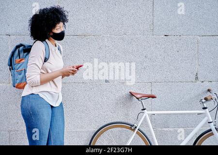 Donna afroamericana con uno smartphone in mano. Donna con maschera protettiva. Andare in bicicletta in città. Foto di alta qualità Foto Stock